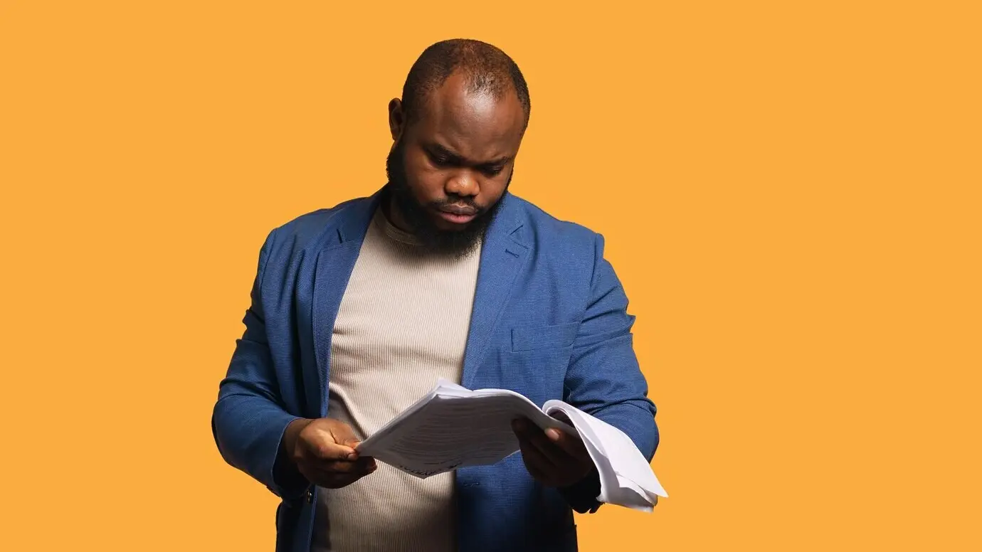 Ein Mann blättert durch Berichtsunterlagen, wirft sie aus Wut weg und fühlt sich überarbeitet.